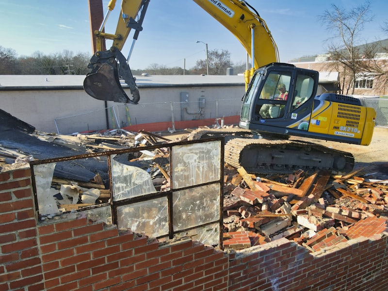 A yellow excavator taking down a masonry wall on a demolition site.