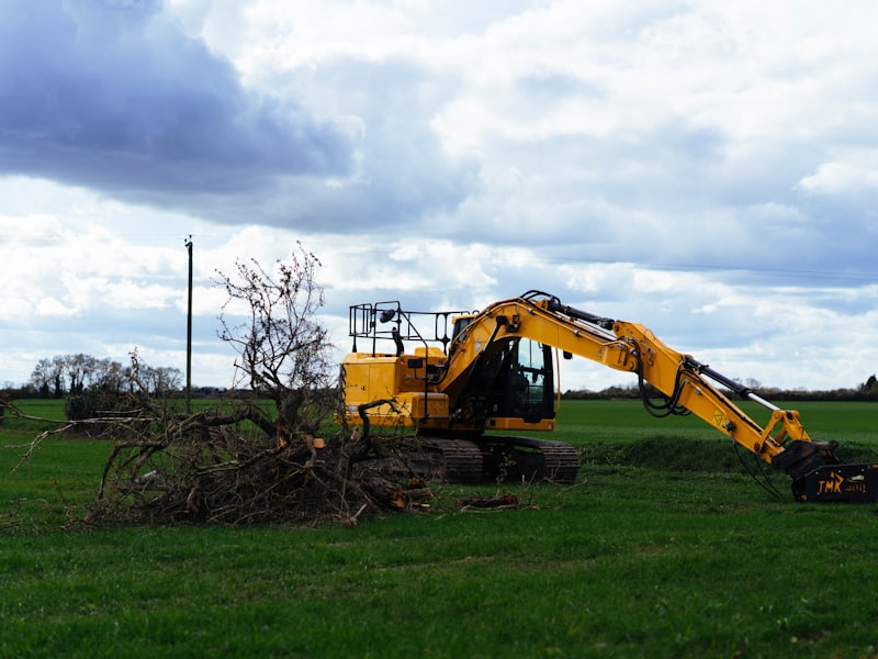 A tracked excavator with a clearing attachment piling brush and tree limbs at the edge of an open field.